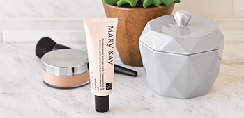 Mary Kay foundation primer sits atop a marble bathroom countertop alongside mineral powder foundation, a makeup brush, gray beauty container and succulent plant.