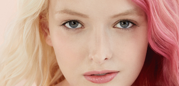 A Caucasian Mary Kay model with half pink, half blonde hair looks confidently at the camera with her hands under her chin.