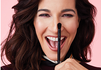 A brunette Latina model smiles while holding a Mary Kay makeup brush in front of her face.